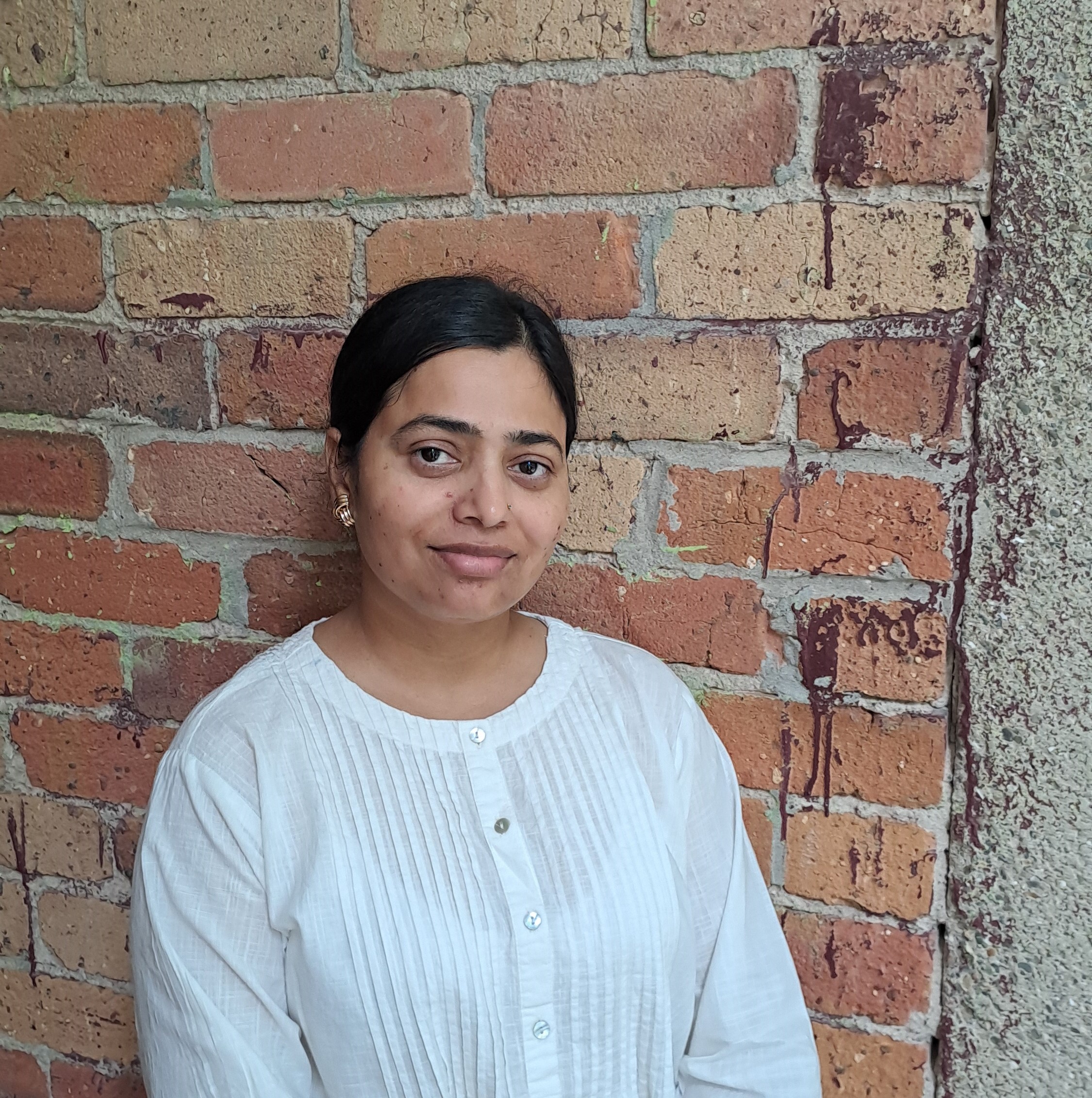 Image of Nilofar standing in front of a brick wall. She is wearing a white shirt and gives a light smile. 