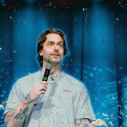 Image of comedian Chris D'Elia with a microphone, mid performance, standing in front of a blue background. 