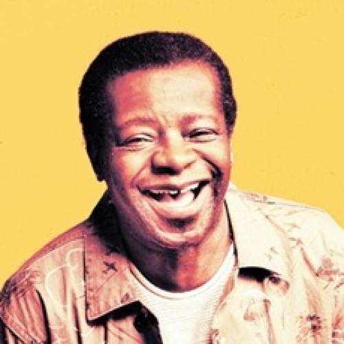 Comedian Stephen K Amos, a black middle aged man with an excellent smile, stands in front of a bright yellow background