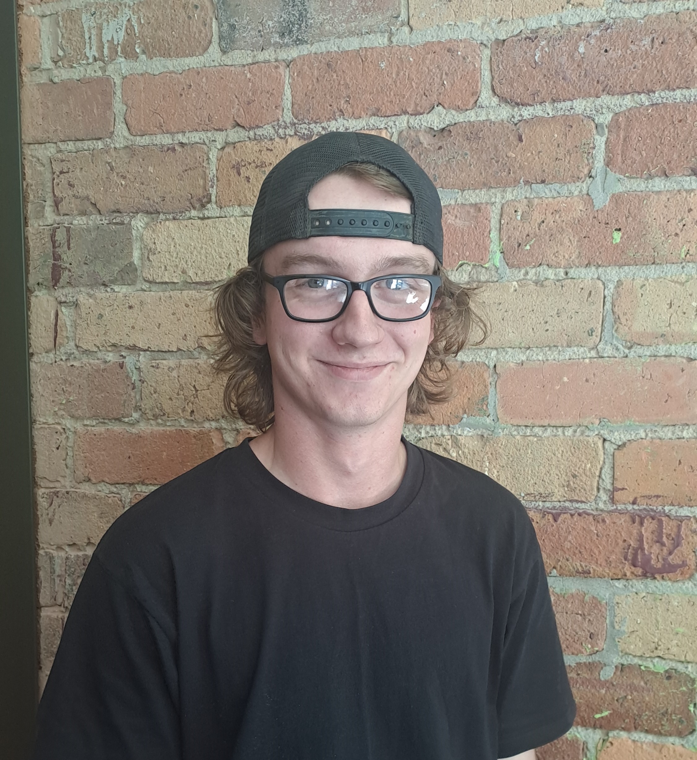 Picture of Dan smiling, standing in front of a brick wall. He wears a black tshirt and cap and glasses