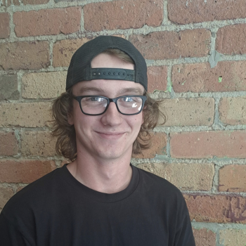 An image of Dan in front of a brick wall. he wears glasses and a backward hat