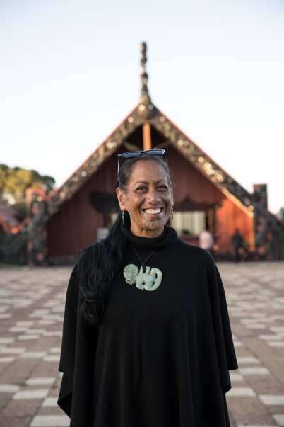 Huia Pihema stands on a marae ātea, the wharenui blured in the background