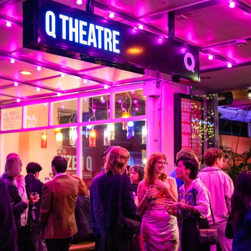 A group of well dressed theatre goers outside of Q Theatres entrance, a large sign reads Q Theatre with pink neon lights surrounding it