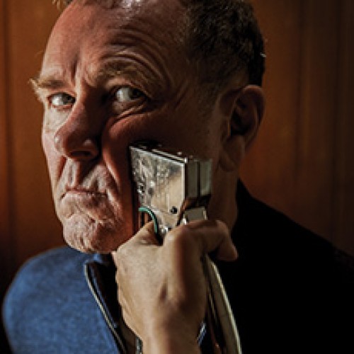 Actor Stephen Lovatt stares deadpan into the camera while an unseen person holds a staple gun to his face