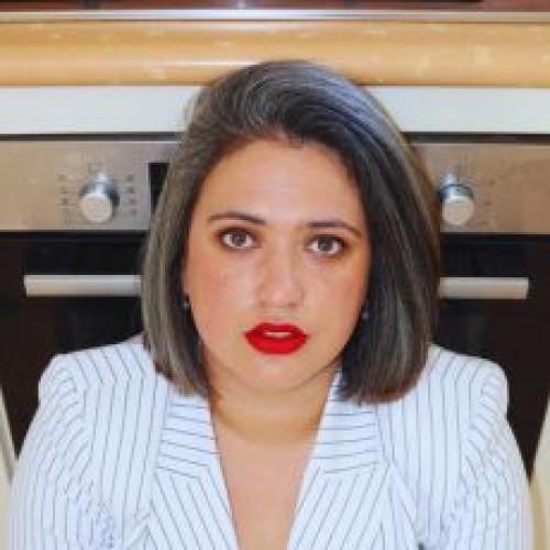 An image of Mo Munn (head and shoulders framing) in front of an oven - she has shoulder length hair, a white jacket with black stripes,and red lipstick. 