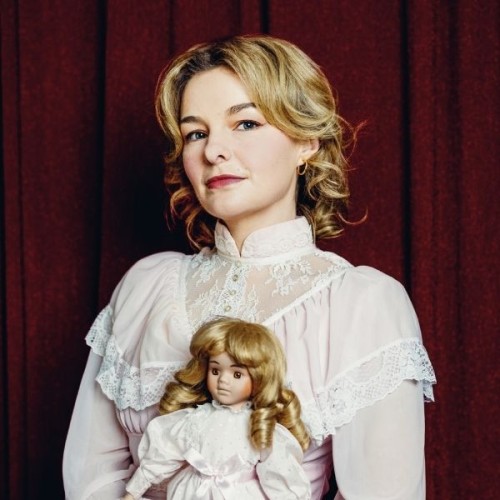Abby Howells stands agaisnt a dark maroon background. She wears her short blonde hair in ringlets and is dressed in a high collared victorian-esque white dress. She looks out with a serious expression. She holds a doll that matches her almost exactly in appearances