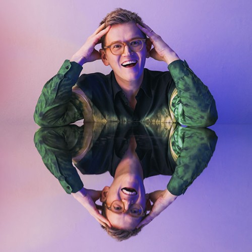 An image of Chris Parker resting his elbows on a reflective surface and his hands agaisnt his head agaisnt a purple gradient background. he is reflected back again underneath himself He wears a green shirt and glasses