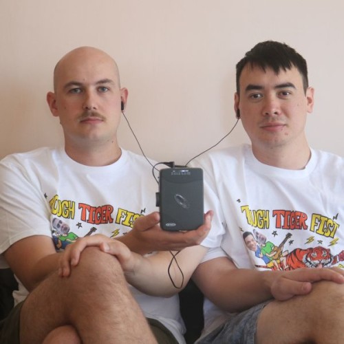 A photo of the two members of Tough Tiger Fist wearing Tough Tiger fist Tshirts sitting listening to a walkman together agaisnt a white background