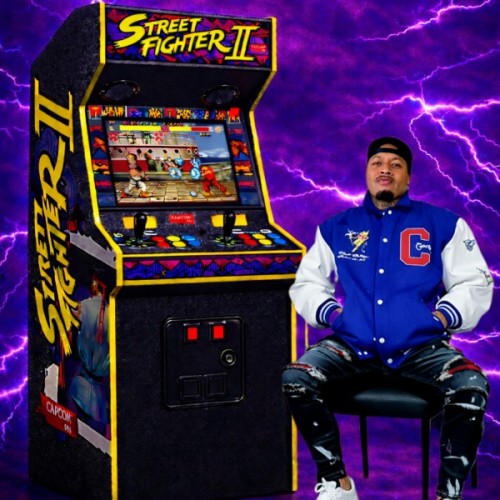 a street fighter 2 pinball machine stand in front of a purple lightening background next to it sits Shady Tapu in a blue letterman jacket and a backwards cap.