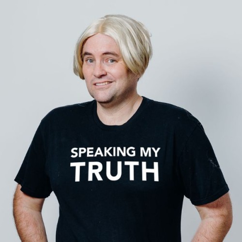 A mid-shot photo of Tom Sainsbury in a black t-shirt that says in white writing 'speaking my truth'. He wears a silver bob wig, and stands agaisnt a grey background. 