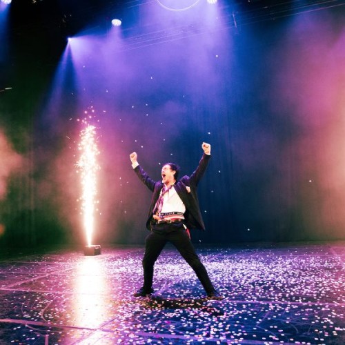 A man stands on a purple stage with fireworks going off beside him
