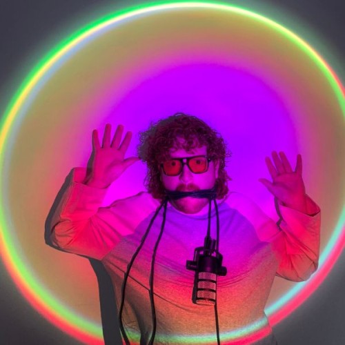 An image of Harrison keefe up agaisnt a wall in a purple tinted spotlight, hands up, mouth taped, glasses on, mic drapped round his shoulders