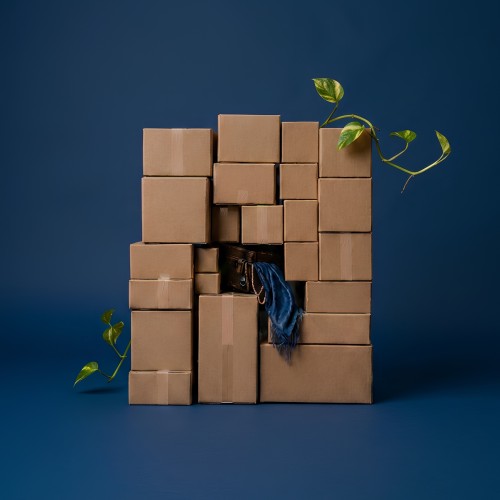 A pile of brown paper cardboard boxes photographed against a blue wall. One box is open and it's contents of fabric and personal items spill out.