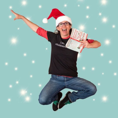 Author R.A. Spratt wearing blue jeans, a black t-shirt, and a red santa hat in a jumping pose holding a book. They are smiling and joyful. The background is pale blue with white snowflakes.