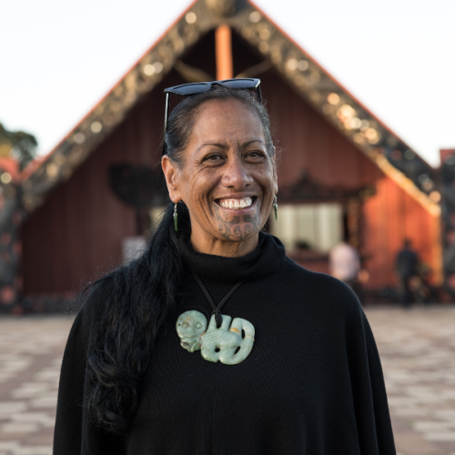Photo of Huia Pihema, a wharenui in the distance behind her